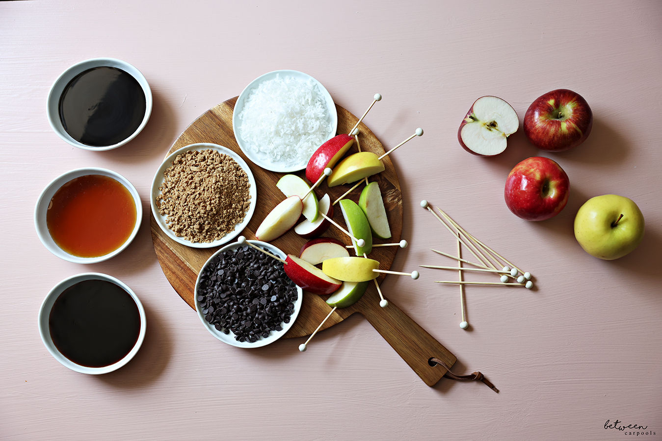 Skewered apple spears, and toppings in shallow bowls, on a wooden board. More toppings, skewers, and a variety of apples, nearby.