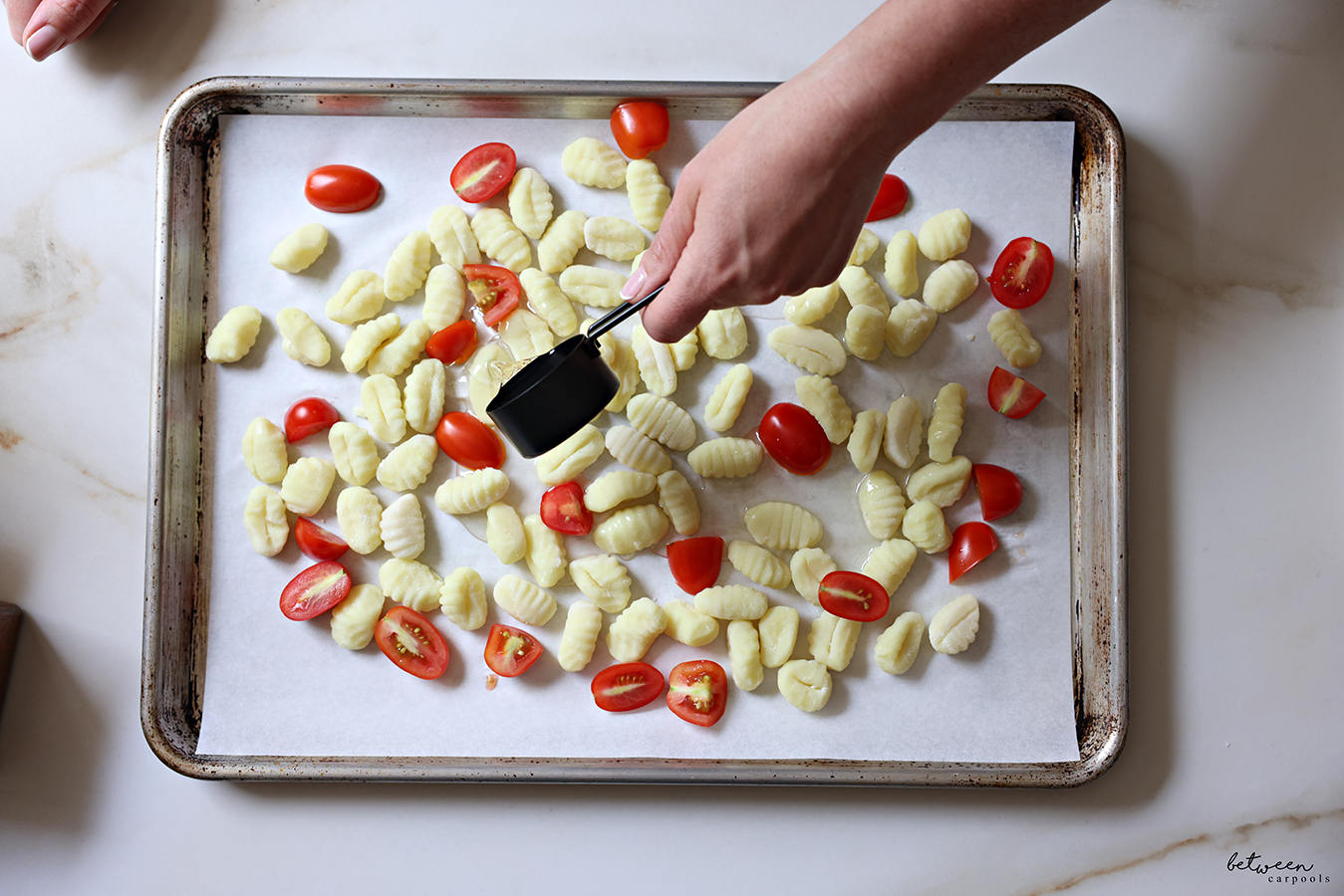 Adding oil to a parchment lined baking sheet with gnocchi and halved grape tomatoes.