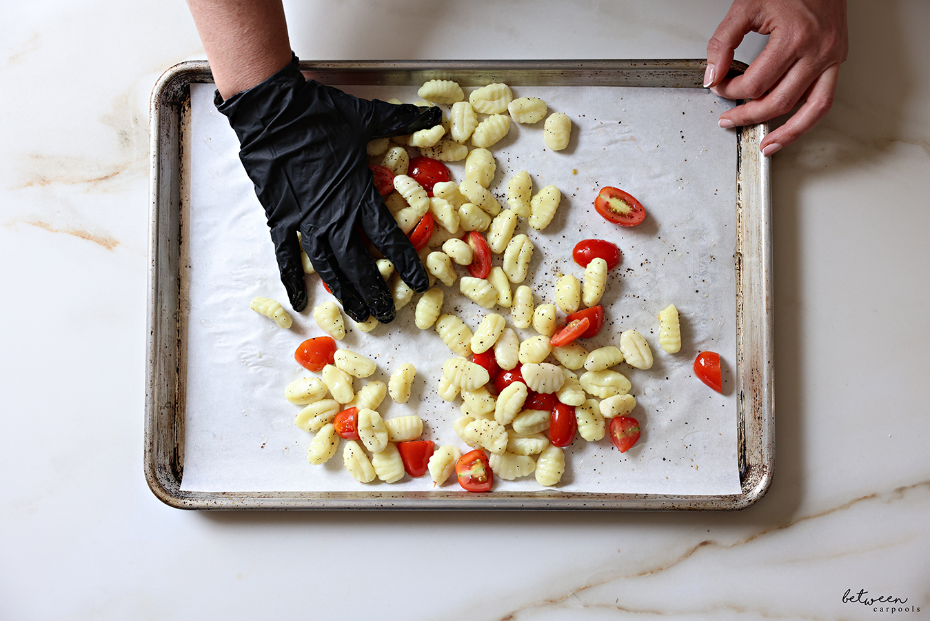 Tossing gnocchi and halved grape tomatoes with oil salt and pepper on a parchment lined baking sheet.