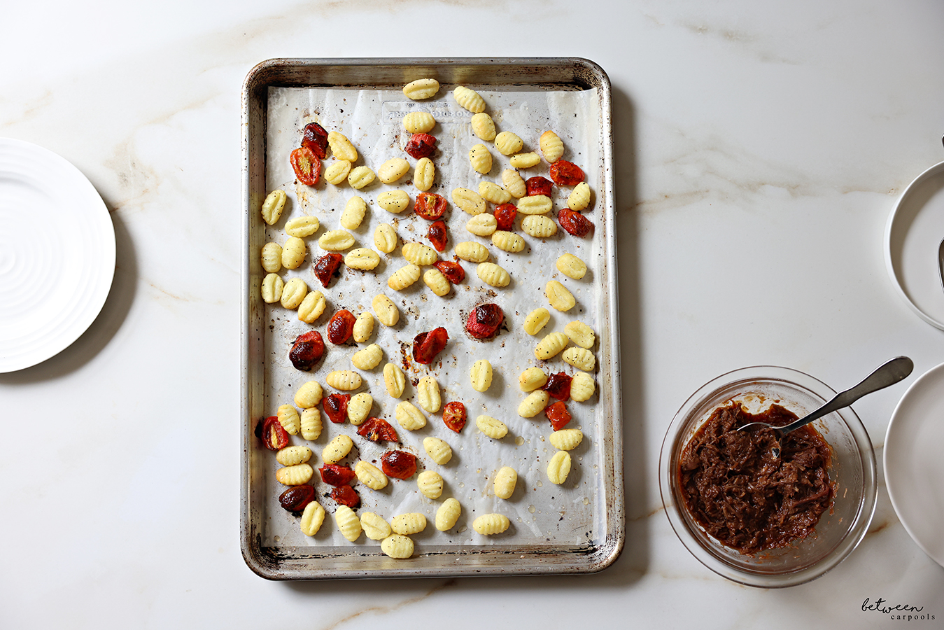 Baked gnocchi and grape tomatoes on a parchment lined baking sheet. Shredded beef in a small glass mixing bowl.